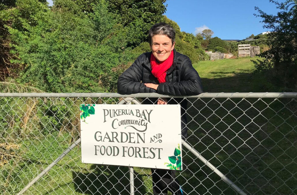 Jane smiling standing at a gate with a hand painted sign that says Pukerua Bay Community Garden and Food Forest