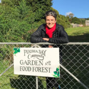 BLOG Community Garden gate Jane smiling standing at a gate with a hand painted sign that says Pukerua Bay Community Garden and Food Forest