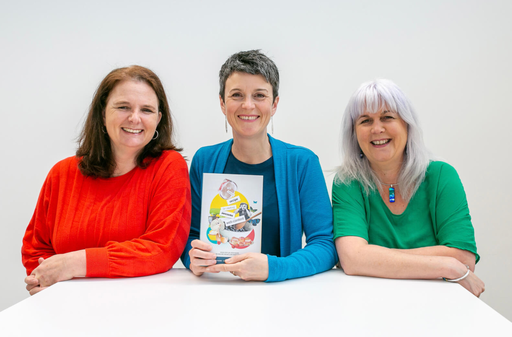 3 smiling women sat at a white table. The middle woman is holding a book.