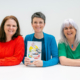 3 smiling women sat at a white table. The middle woman is holding a book.