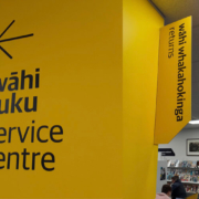 Yellow wall with a left pointing arrow with the words wahi tutu service centre with library shelves and people reading in the background