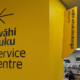 Yellow wall with a left pointing arrow with the words wahi tutu service centre with library shelves and people reading in the background